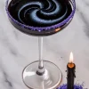 Halloween cocktail with a hypnotic black and blue swirl served in a coupe glass with a sparkly purple sugar rim, accompanied by a burning black candle resting in purple sugar on a marble counter.