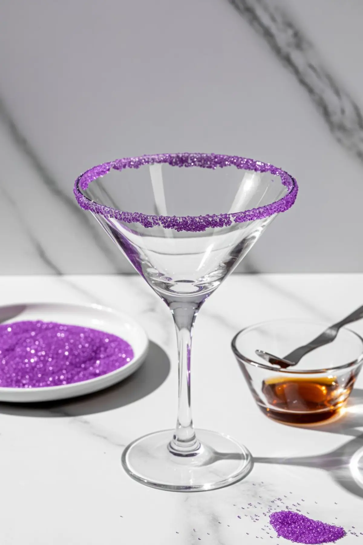 Empty martini glass with a thick rim of purple sanding sugar beside a small bowl of amber liquid and a plate of glittery sugar, set on a white marble surface.
