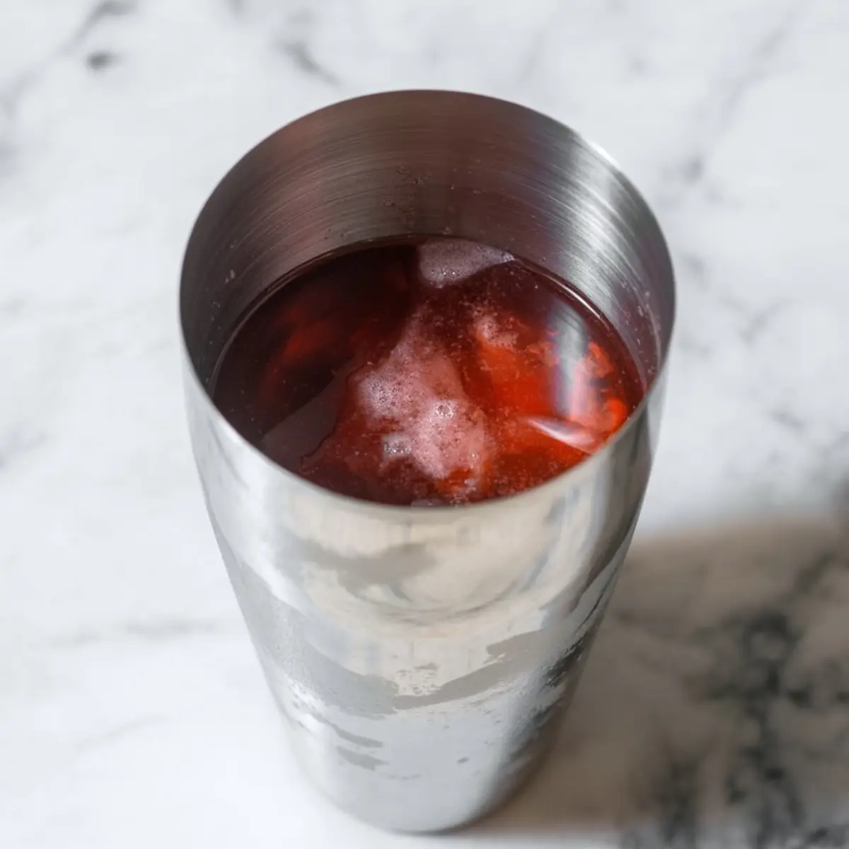Stainless steel cocktail shaker filled with red Halloween margarita mix over ice, placed on a marble countertop.