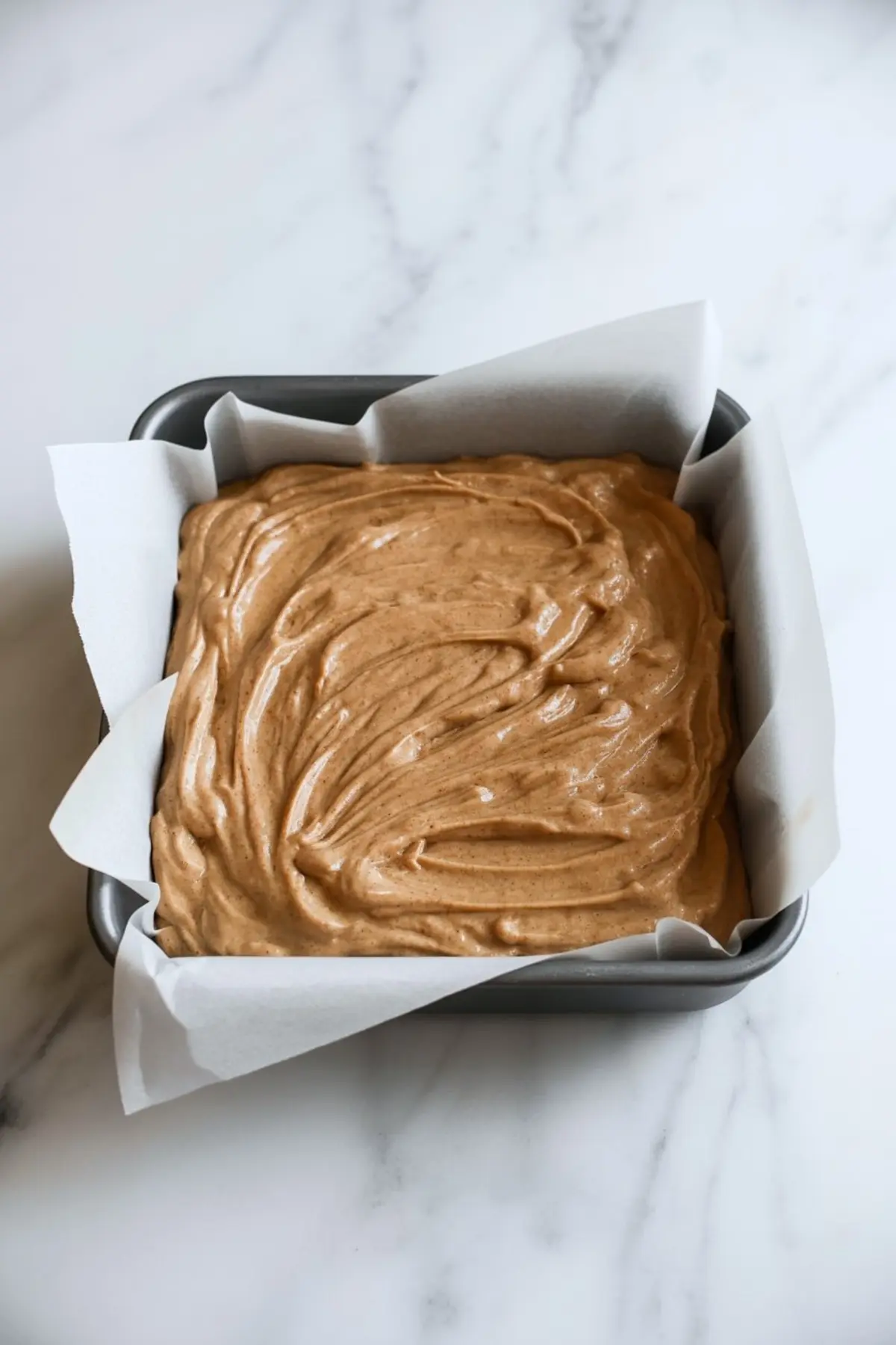 Square baking pan lined with parchment paper filled with smooth brown snickerdoodle bar batter, ready for baking.