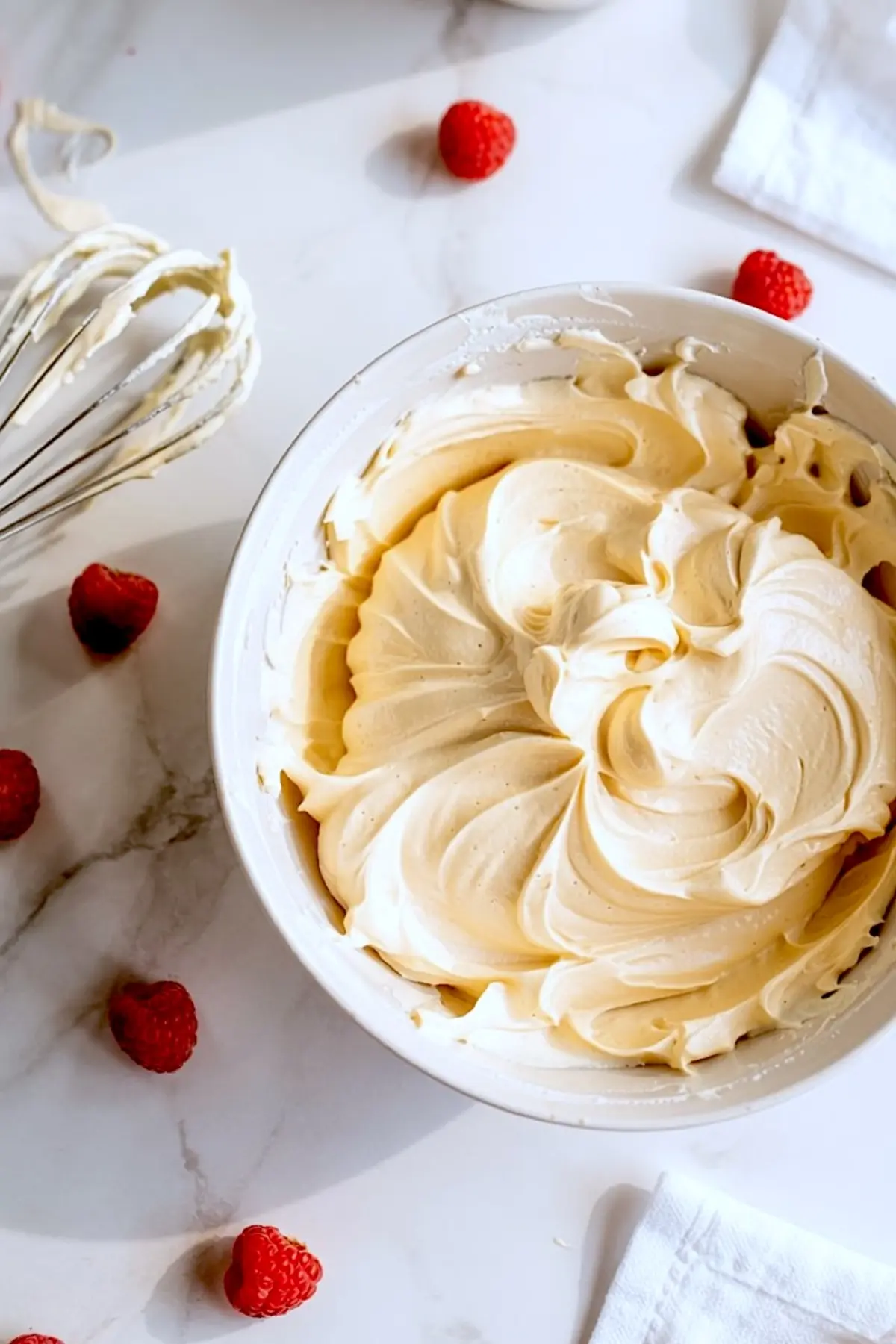 Swirled whipped buttercream frosting in a white bowl surrounded by fresh raspberries and a wire whisk on a white marble background.