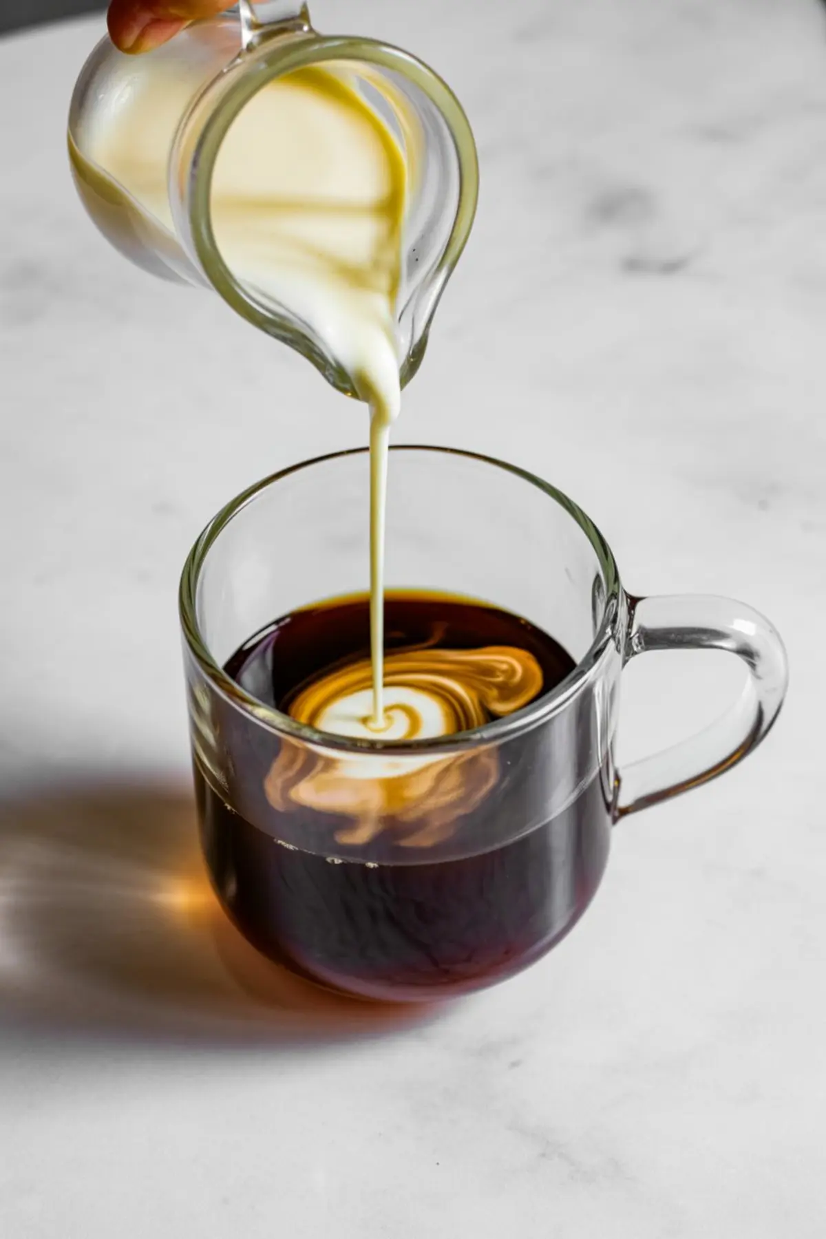 Sweetened cream being poured into a glass mug of black coffee, forming a swirling pattern on the surface, with a soft gradient of light and dark tones.