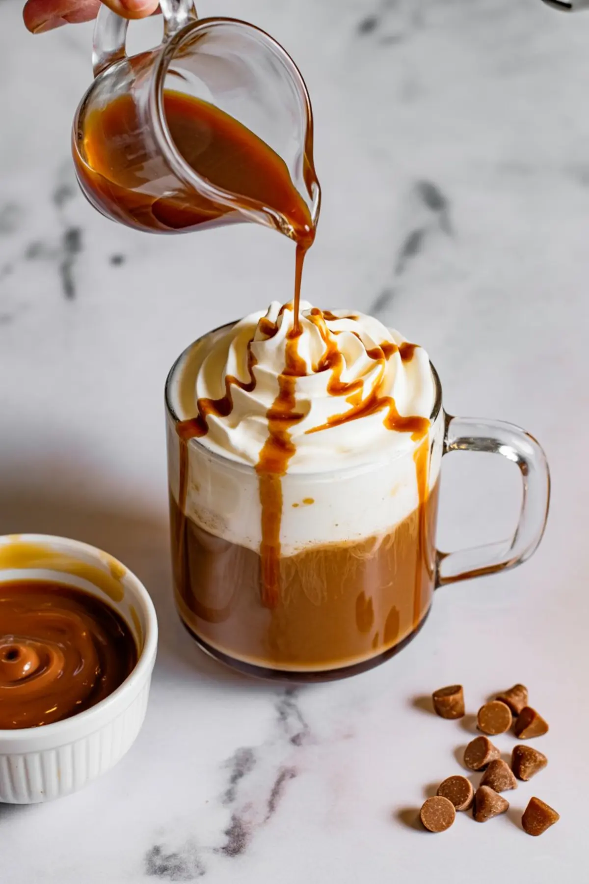 Caramel sauce being poured from a glass pitcher onto whipped cream over a glass mug of butterscotch coffee, with dulce de leche in a ramekin and butterscotch chips on the side.