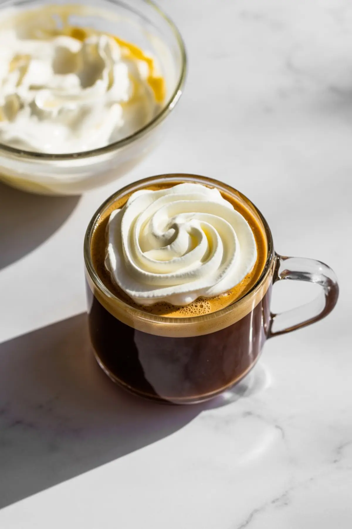 Glass mug of espresso topped with a thick swirl of whipped cream, placed beside a glass bowl of cream on a sunlit marble surface.