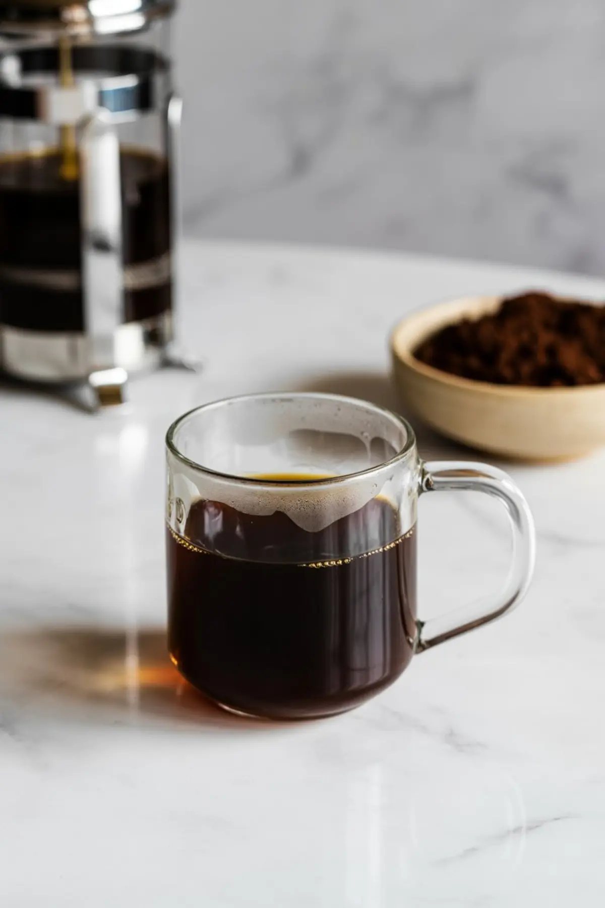 Freshly brewed black coffee served in a clear glass mug with a French press and a bowl of ground coffee in the background on a marble countertop.