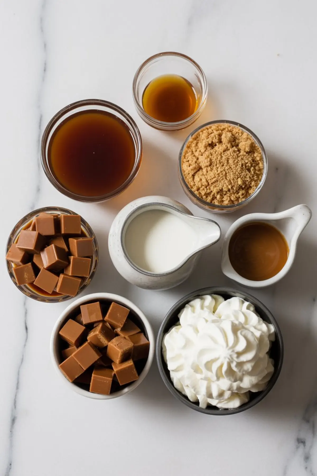 Flat lay of butterscotch coffee ingredients including caramel cubes, brown sugar, whipped cream, vanilla extract, milk, and caramel sauce, arranged in glass and ceramic bowls on a marble background.