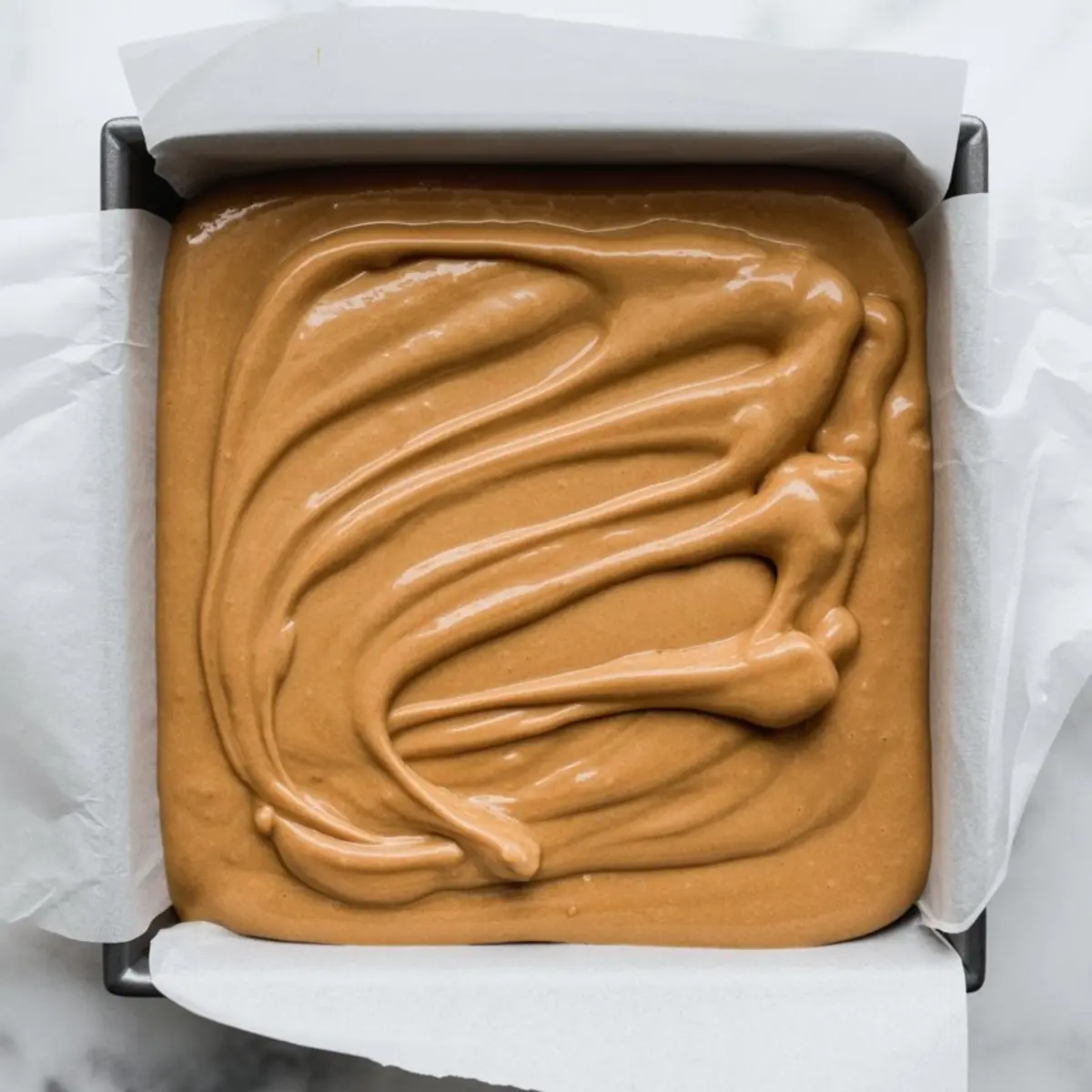 Square pan filled with freshly poured butterscotch fudge mixture in smooth swirls, lined with parchment paper and ready for chilling before cutting into squares.
