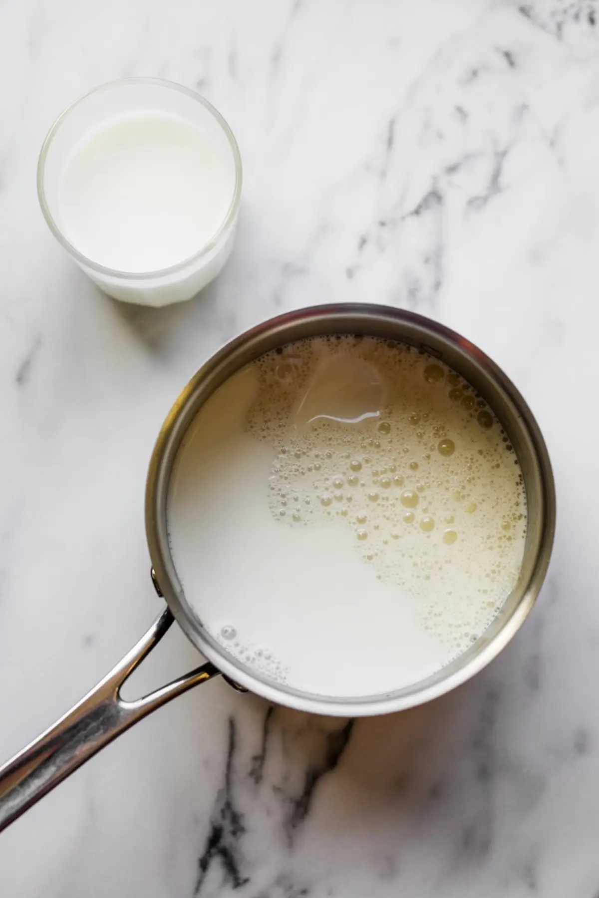 Saucepan with steaming milk next to a glass of milk on a white marble counter, part of the cooking process for butterscotch pudding.