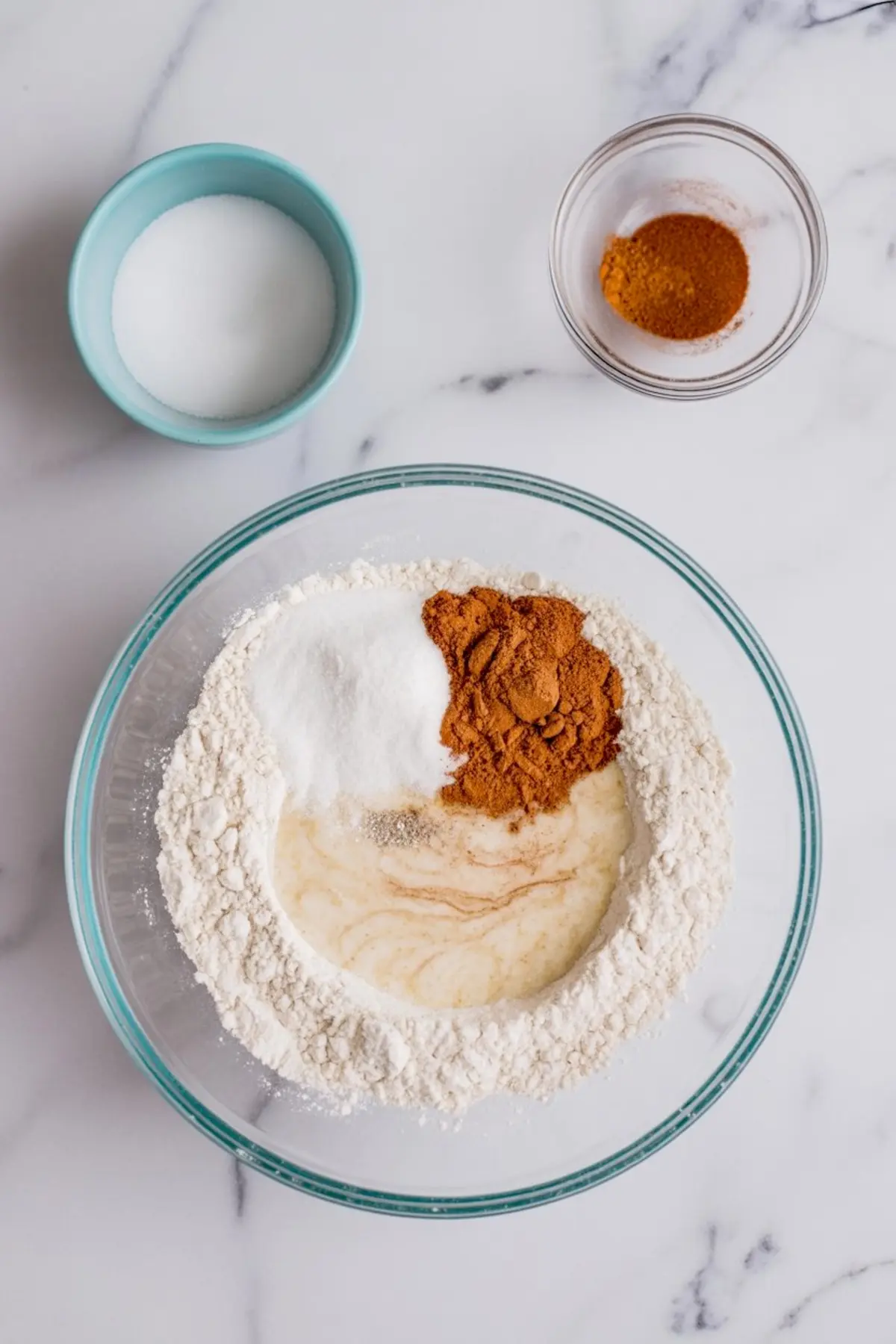 Top view of dry baking ingredients in a glass bowl, including flour, granulated sugar, cinnamon, nutmeg, and vanilla extract.