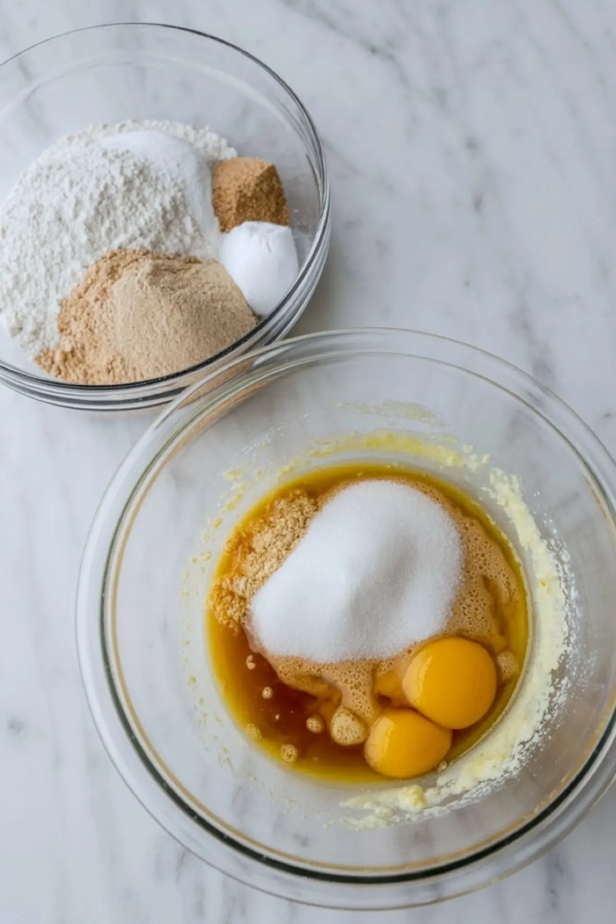 Two glass mixing bowls filled with cookie ingredients, including flour, baking soda, brown sugar, white sugar, eggs, and vanilla extract, set on a marble surface.