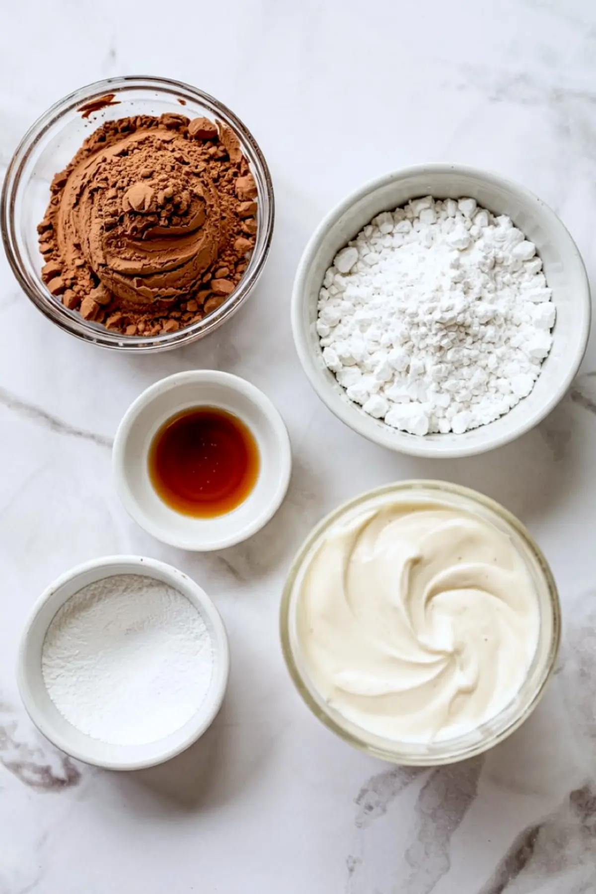 Flat lay of individual bowls holding unsweetened cocoa powder, powdered sugar, baking soda, vanilla extract, and whipped topping on a light marble background.
