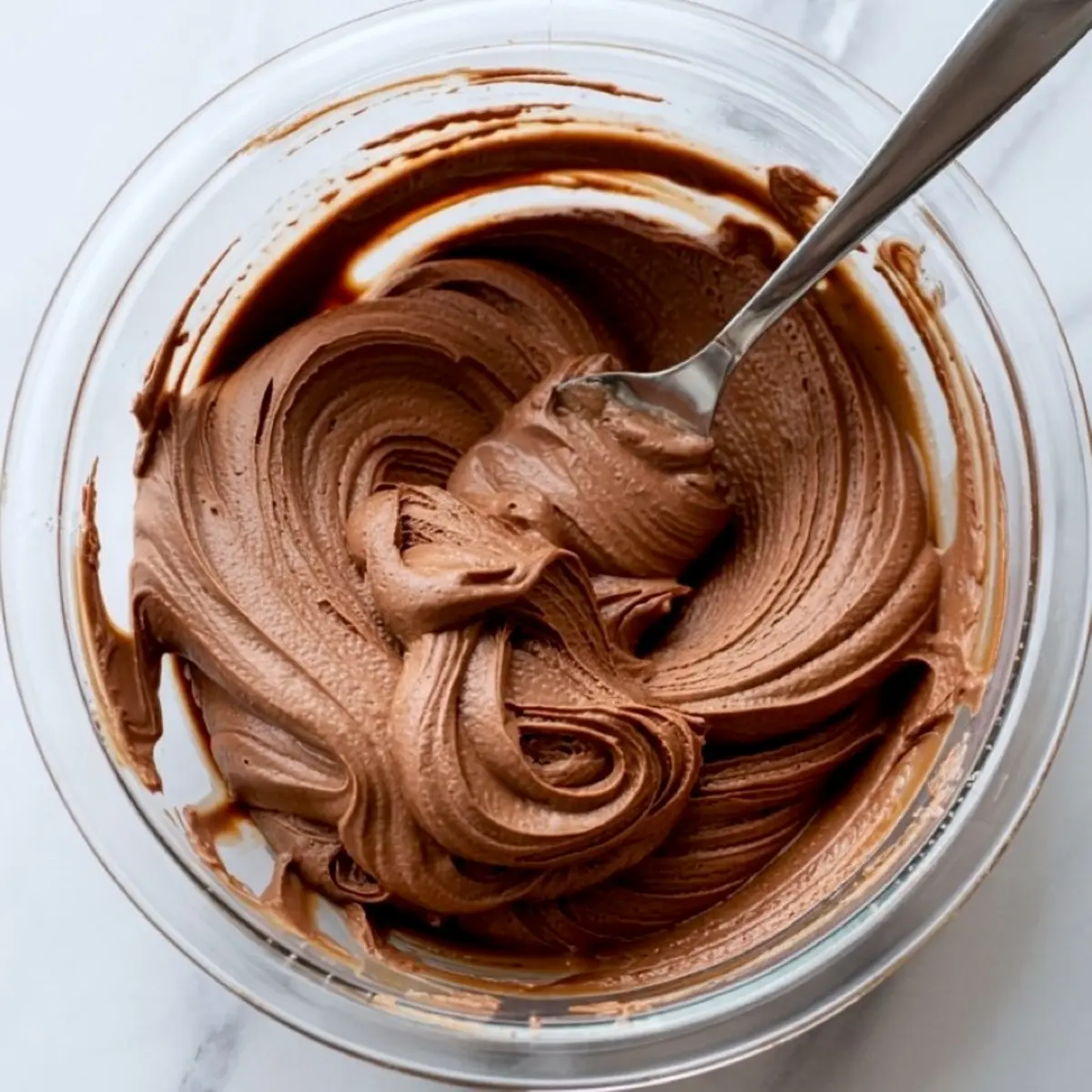 Thick chocolate pudding frosting mixture being stirred with a spoon in a glass bowl.
