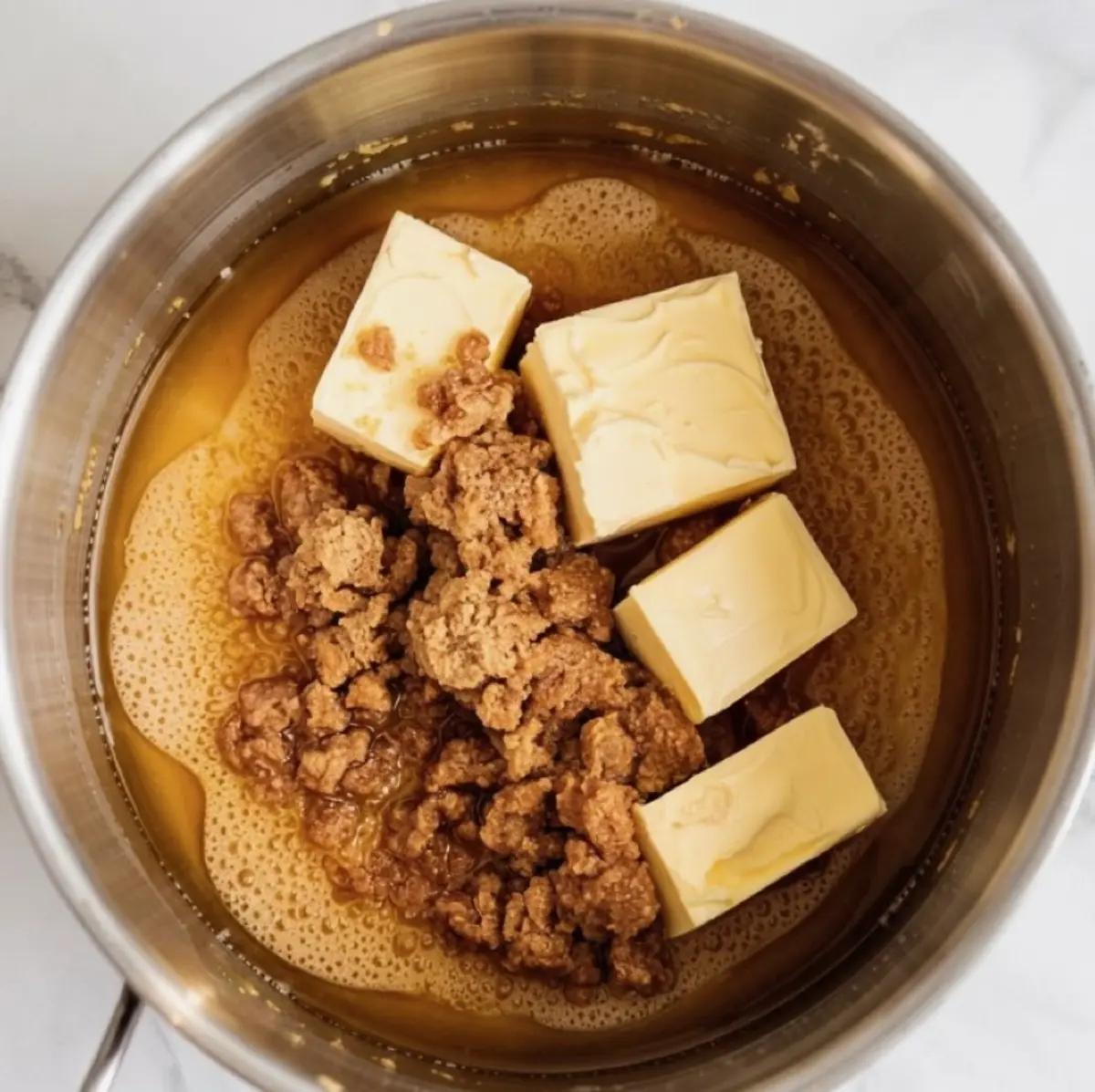 Overhead shot of solid butter cubes and crumbled brown sugar placed over hot, partially melted caramel mixture in a metal saucepan.