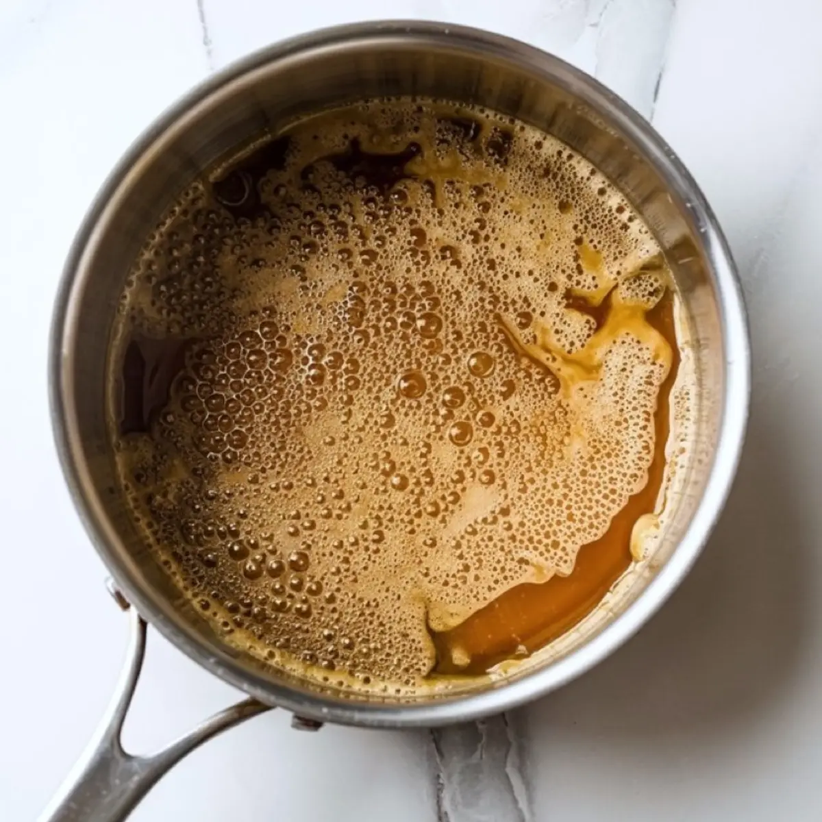 Top view of bubbling golden-brown toffee syrup cooking in a stainless steel saucepan on a white marble background.