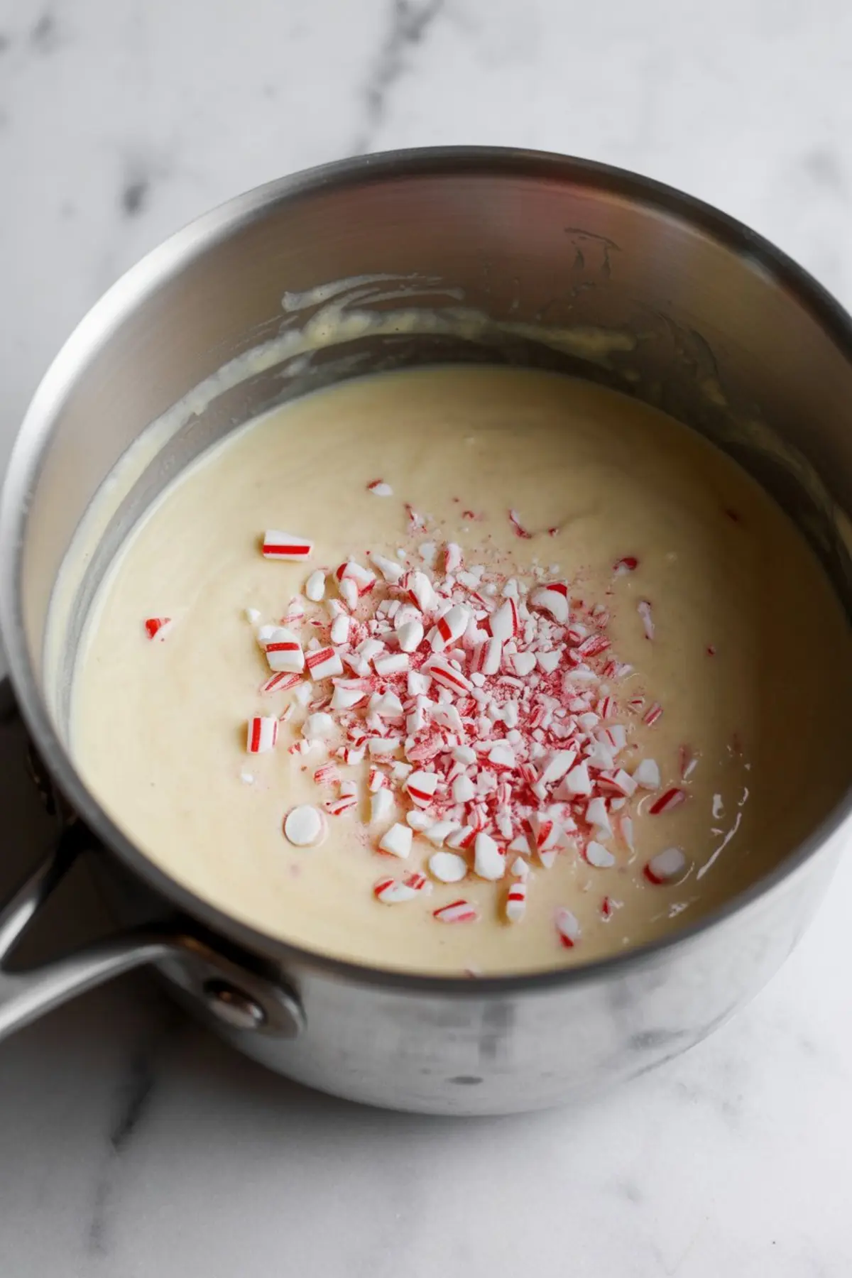 Smooth white chocolate fudge mixture in a saucepan with crushed candy canes added on top, showing the preparation process for peppermint Christmas fudge.