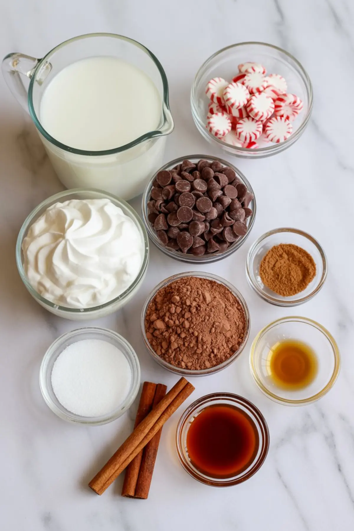 Flat lay of hot chocolate ingredients on a marble surface, including milk, whipped cream, semi-sweet chocolate chips, cocoa powder, granulated sugar, peppermint candies, cinnamon sticks, vanilla extract, and peppermint extract.
