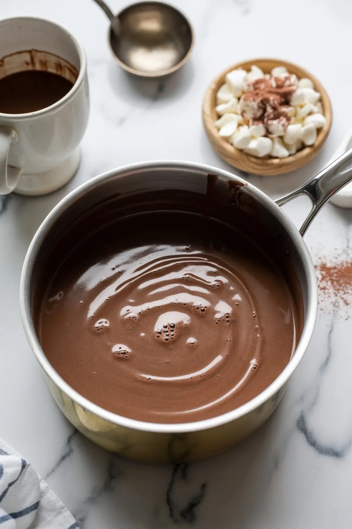 Overhead view of rich homemade hot chocolate in a saucepan, surrounded by a white mug with chocolate residue, a ladle, and a small wooden bowl of mini marshmallows topped with cocoa powder.
