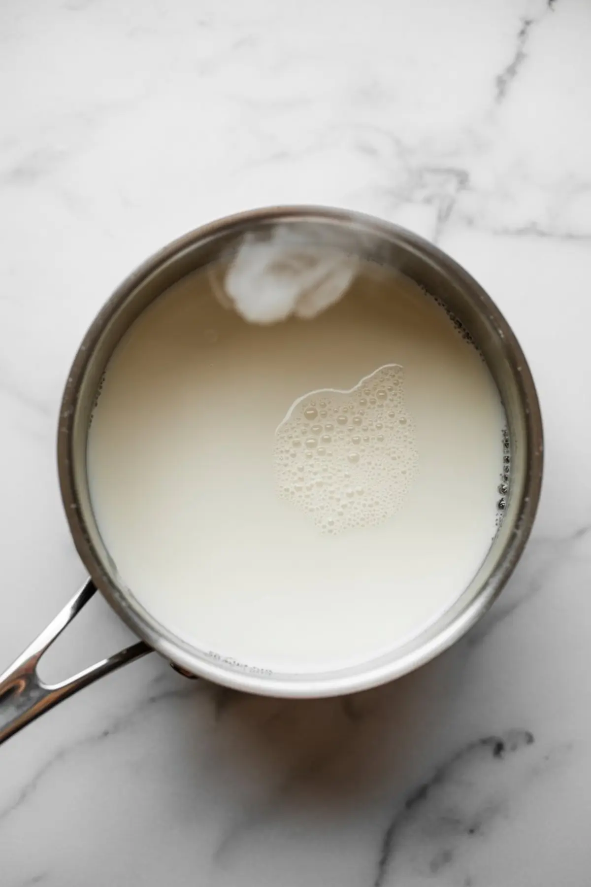 Top view of milk heating in a stainless steel saucepan over a marble countertop, with light foam forming on the surface.
