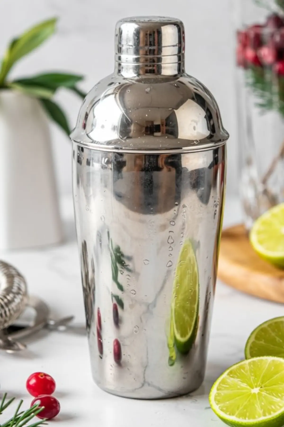 Stainless steel cocktail shaker with condensation droplets, placed on a marble surface next to sliced lime, cranberries, and bar tools for holiday drink preparation.
