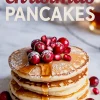 Stack of golden pancakes drizzled with maple syrup, topped with cranberries and pomegranate seeds, and dusted with powdered sugar, with “Christmas Pancakes” text above.