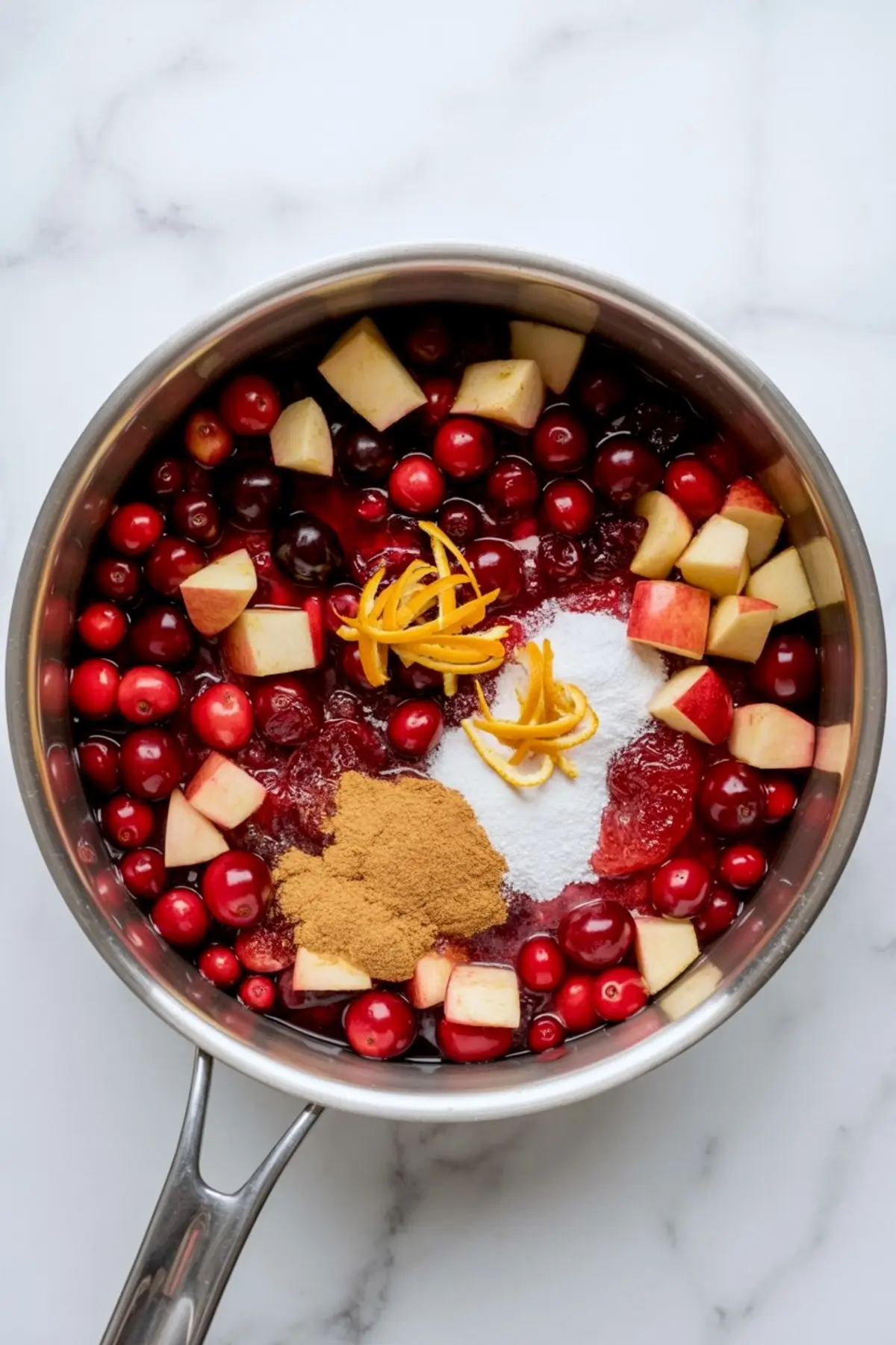 Saucepan filled with fresh cranberries, chopped apples, orange zest, cinnamon, sugar, and cranberry sauce on a white marble background.
