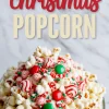 A close-up of a festive bowl of Christmas popcorn coated in white chocolate and topped with red and green candies, peppermint swirls, and holiday sprinkles, with a "Christmas Popcorn" text overlay above.
