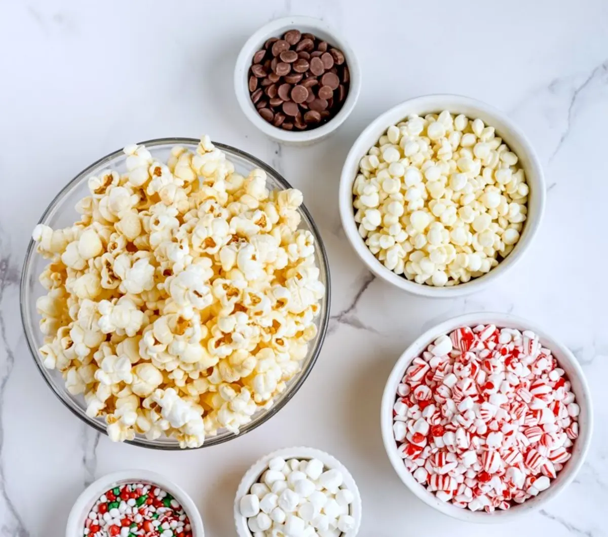 A flat lay of plain popcorn in a glass bowl surrounded by bowls of toppings including white chocolate chips, milk chocolate chips, crushed candy canes, mini marshmallows, and festive red and green sprinkles.