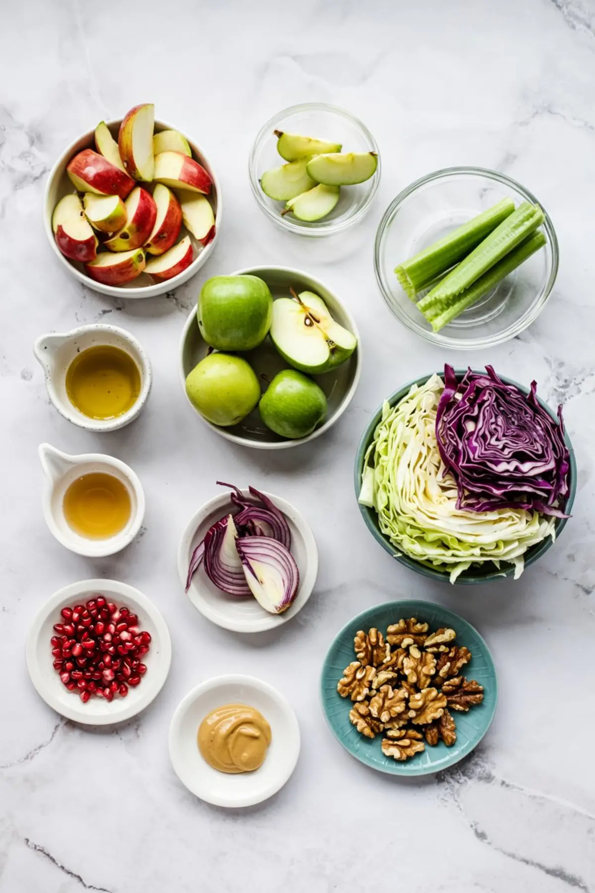 Flat lay of fresh Christmas salad ingredients including red and green apple slices, celery stalks, green and purple cabbage halves, red onion wedges, pomegranate seeds, Dijon mustard, olive oil, apple cider vinegar, and walnut halves on small plates and bowls.
