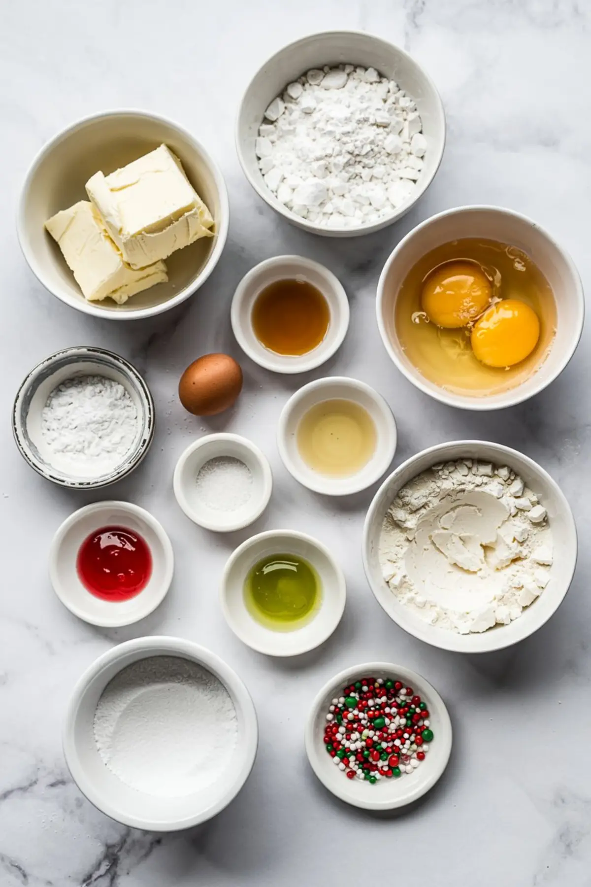 Overhead view of separated Christmas sugar cookie bar ingredients in white bowls, including butter, flour, powdered sugar, eggs, vanilla extract, red and green food coloring, and festive sprinkles.