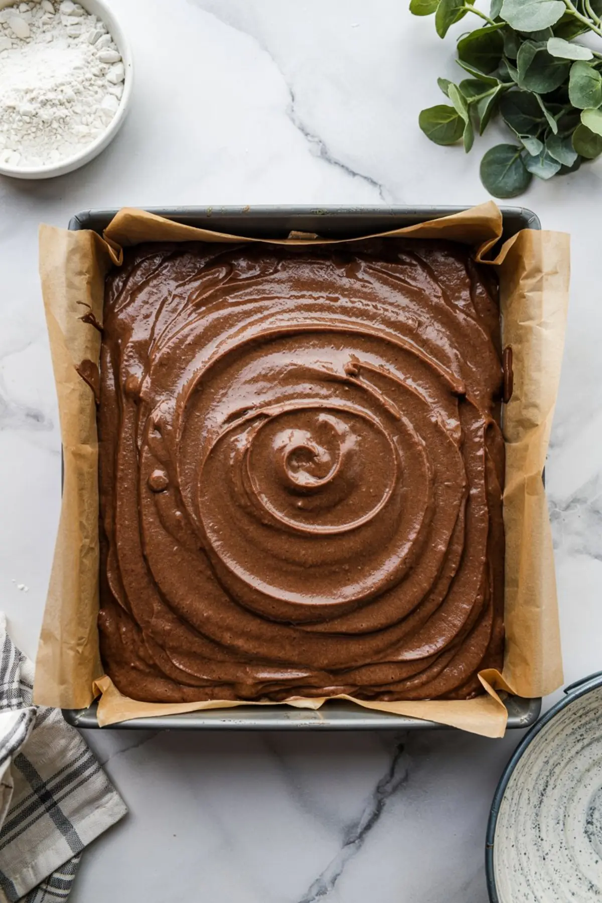 Chocolate brownie batter swirled smoothly in a parchment-lined square baking pan, ready for baking on a white marble surface with flour and greenery nearby.
