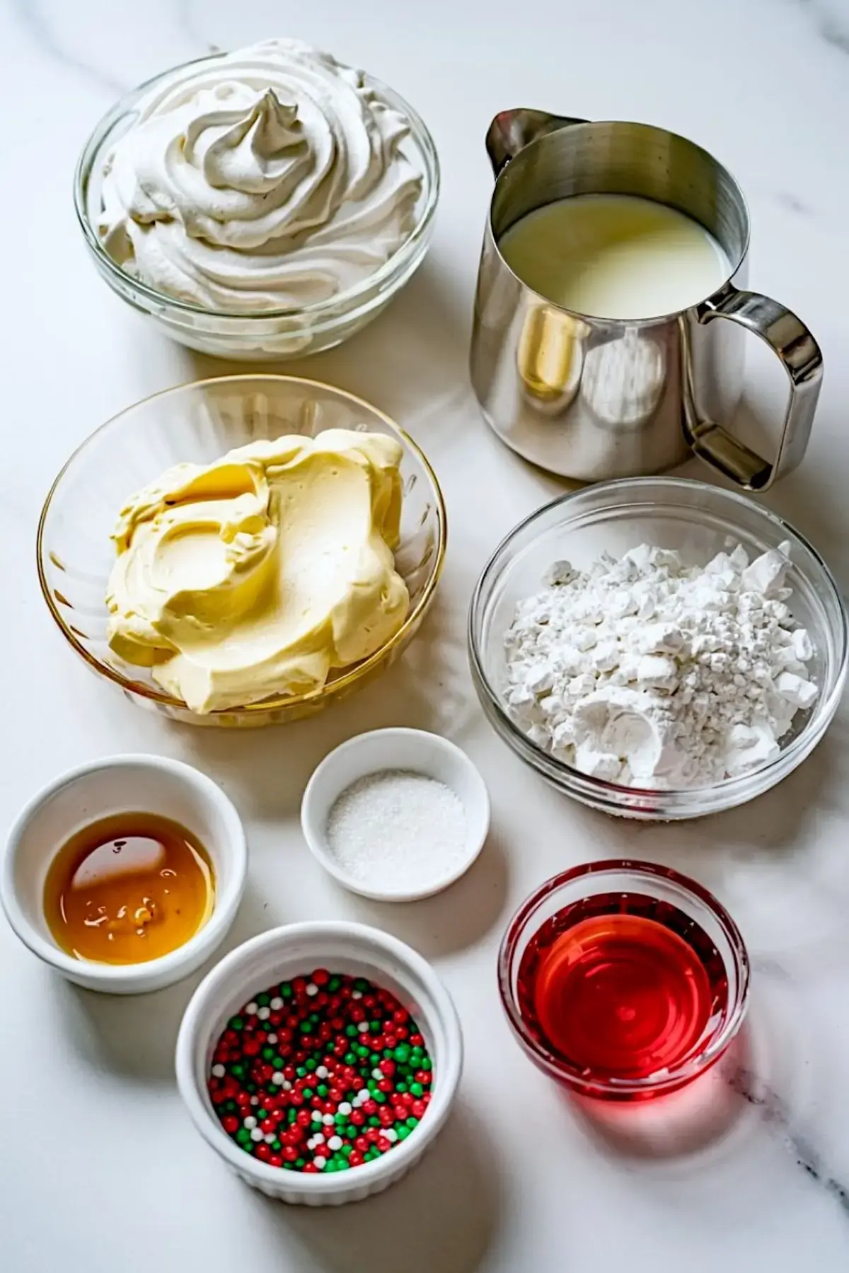 Flat lay of Christmas cake dip ingredients arranged in clear and white bowls, including whipped topping, cream cheese, powdered sugar, red syrup, vanilla extract, white sugar, milk, and red-green holiday sprinkles on a white marble background.
