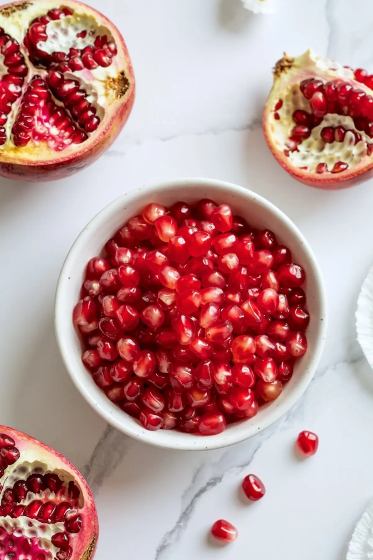 Close-up of a white bowl filled with fresh pomegranate seeds surrounded by halved pomegranates on a marble surface, ideal for holiday fruit platter ideas and healthy snack presentations.
