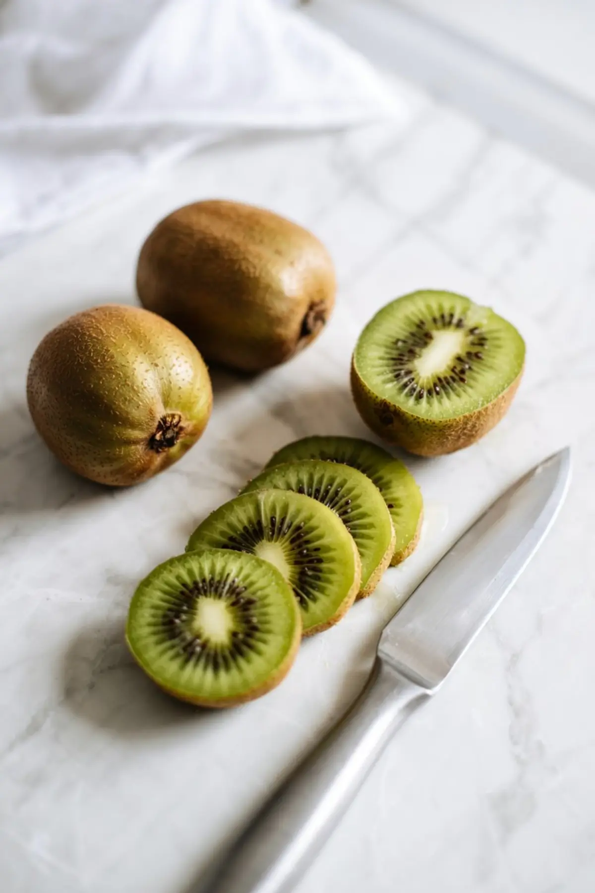Whole and sliced kiwis arranged beside a silver knife on a marble surface, showcasing fresh tropical fruit ideas and ingredients for Christmas fruit salads and smoothie bowls.

