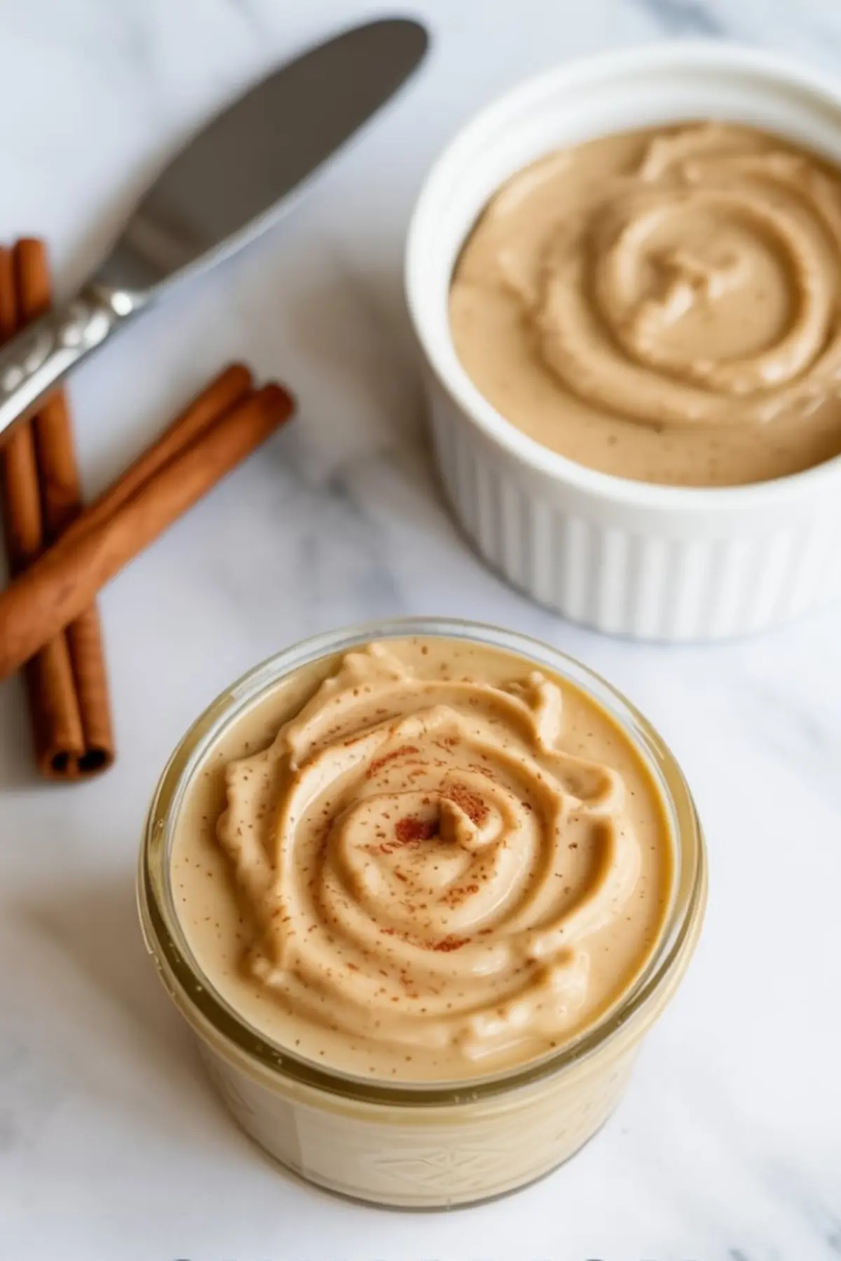 Two small jars of whipped cinnamon butter topped with ground cinnamon, placed beside whole cinnamon sticks and a butter knife on a white marble background.
