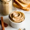 Creamy cinnamon butter in a white ramekin and glass jar, surrounded by cinnamon sticks and slices of toast spread with butter on parchment paper.
