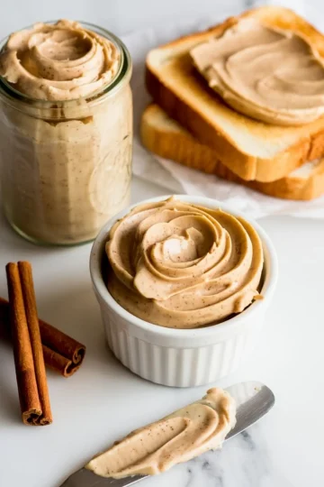 Creamy cinnamon butter in a white ramekin and glass jar, surrounded by cinnamon sticks and slices of toast spread with butter on parchment paper.