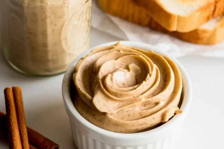 Creamy cinnamon butter in a white ramekin and glass jar, surrounded by cinnamon sticks and slices of toast spread with butter on parchment paper.