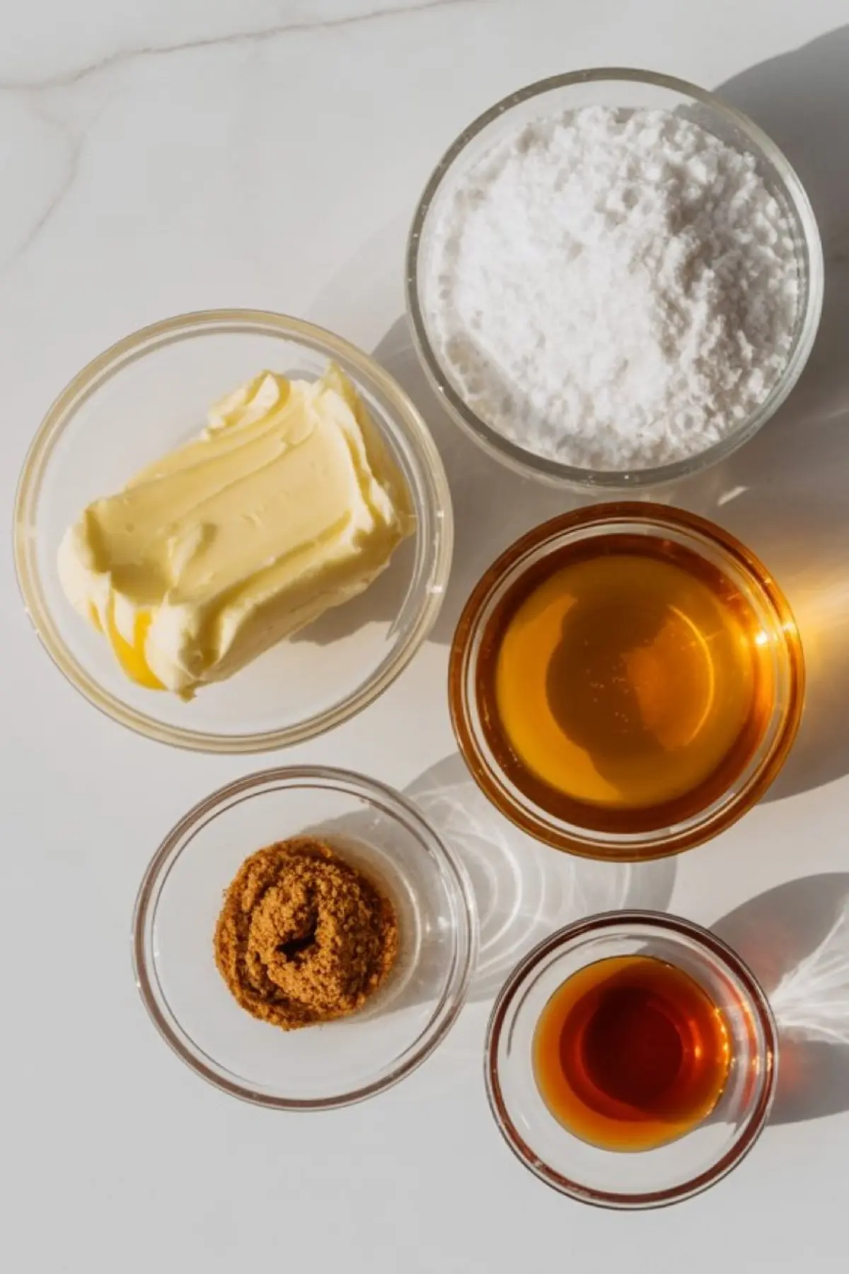 Flat lay of six glass bowls with ingredients for cinnamon butter, including softened butter, powdered sugar, brown sugar, vanilla extract, honey, and cinnamon on a white background.
