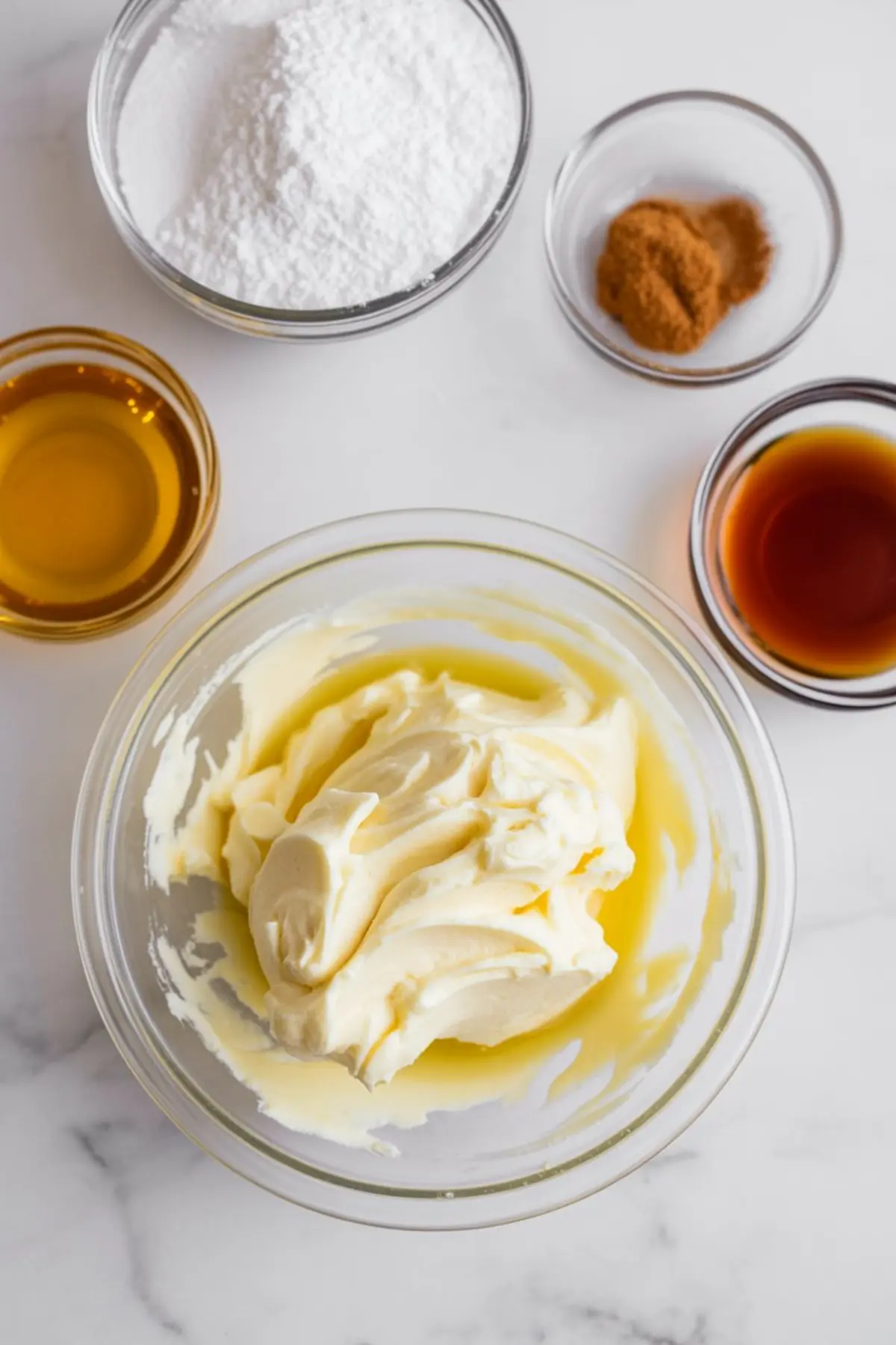 Whipped butter in a clear glass bowl surrounded by ingredients like powdered sugar, cinnamon, honey, and vanilla on a white marble surface.
