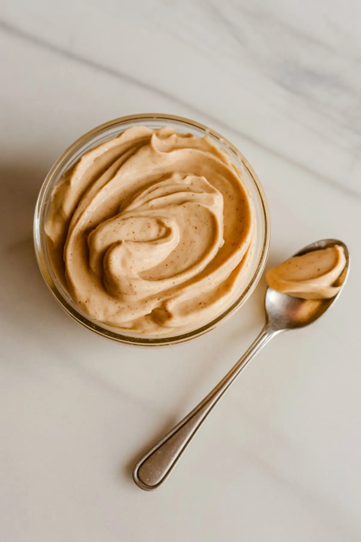 Smooth and creamy cinnamon butter in a glass bowl with a silver spoon holding a dollop on a light-colored background.
