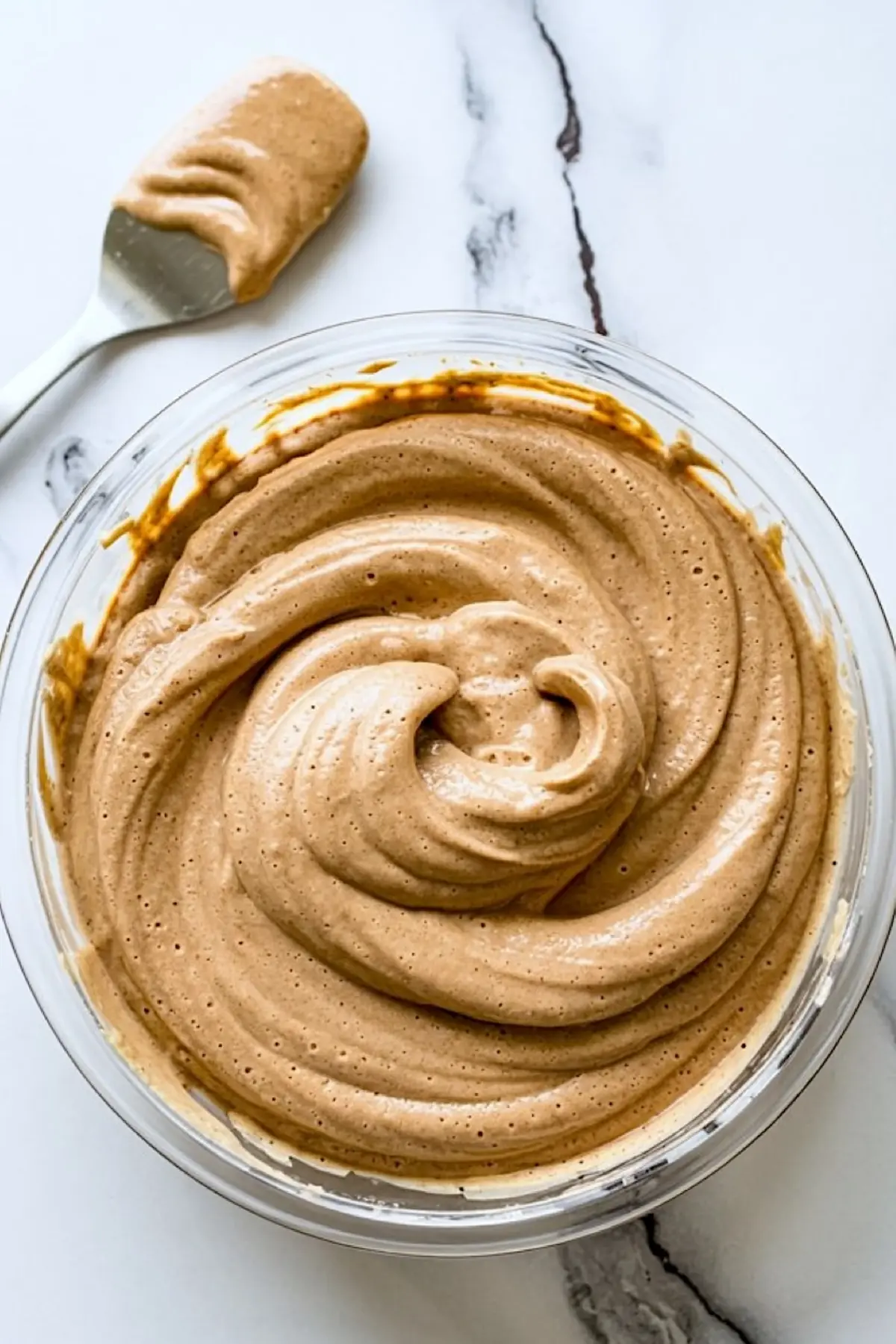 Thick cinnamon pancake batter swirled in a glass bowl with a silicone spatula coated in batter resting beside it on a white marble countertop.