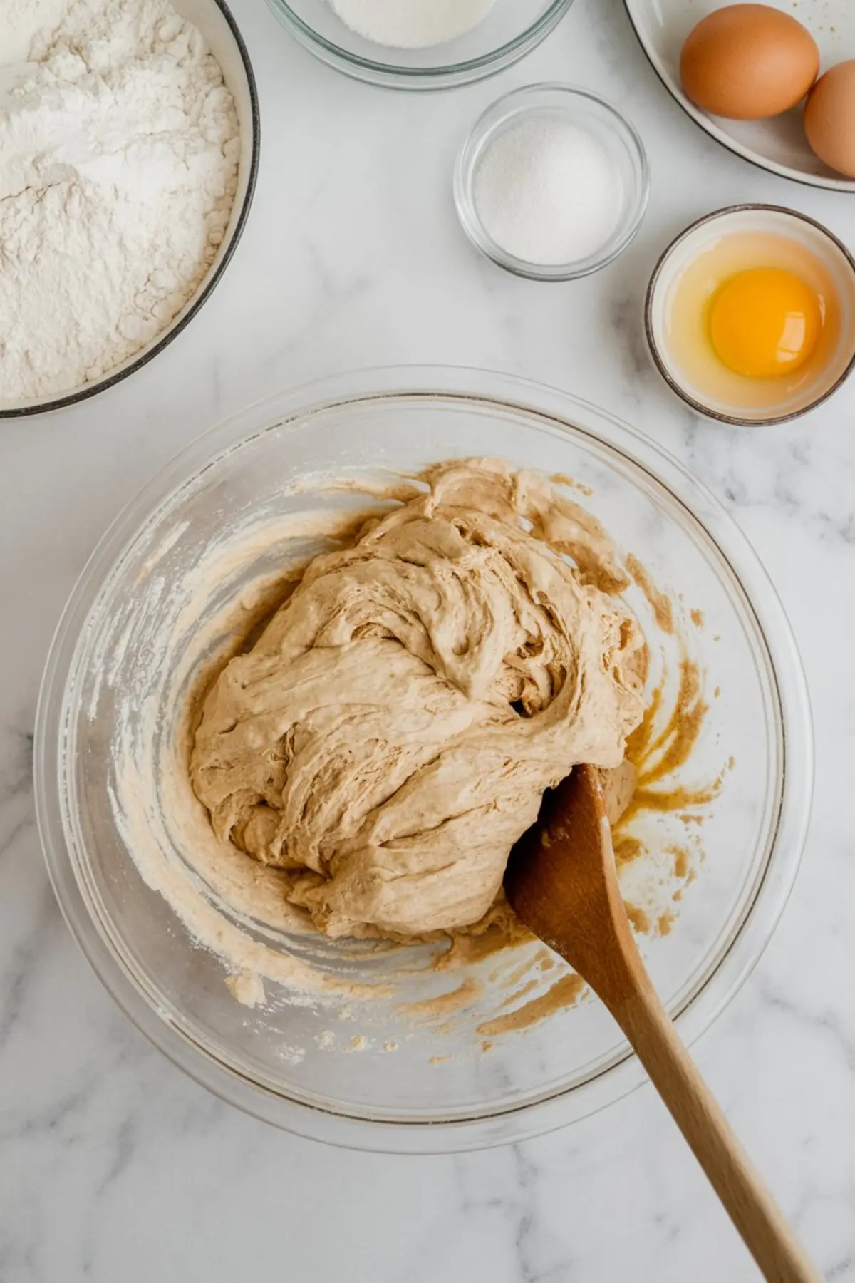 Glass mixing bowl filled with partially mixed cinnamon roll dough, surrounded by baking ingredients including flour, eggs, and sugar on a marble countertop.