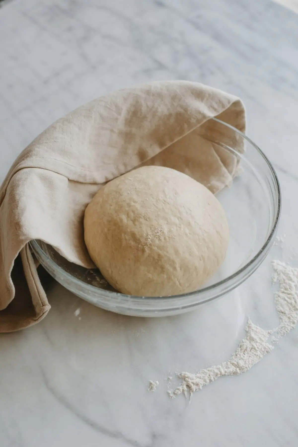 Ball of cinnamon roll dough resting in a glass bowl covered with a beige linen cloth, placed on a floured marble surface.
