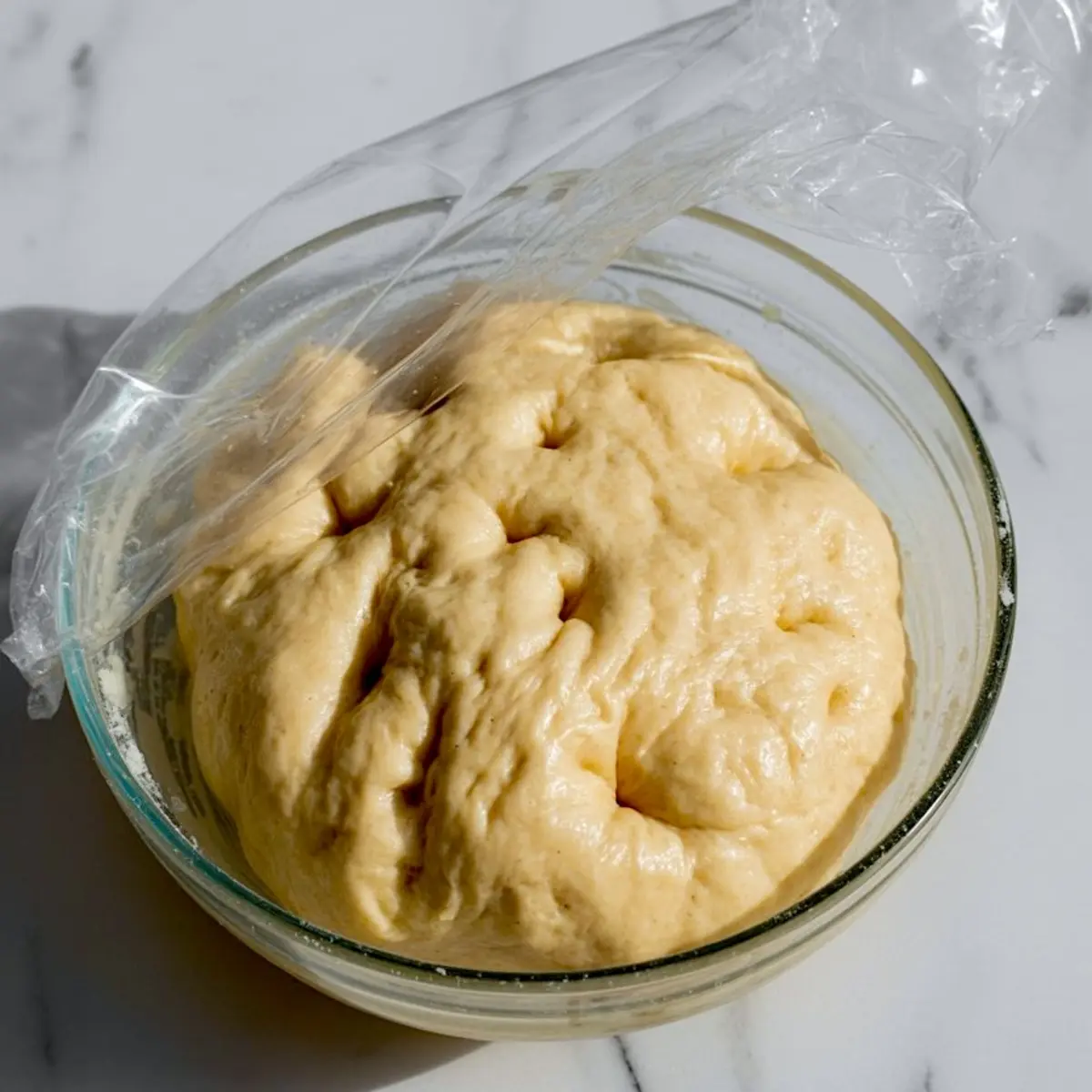 Risen focaccia dough in a glass bowl partially covered with plastic wrap, showing a puffy and well-fermented texture ready for shaping.

