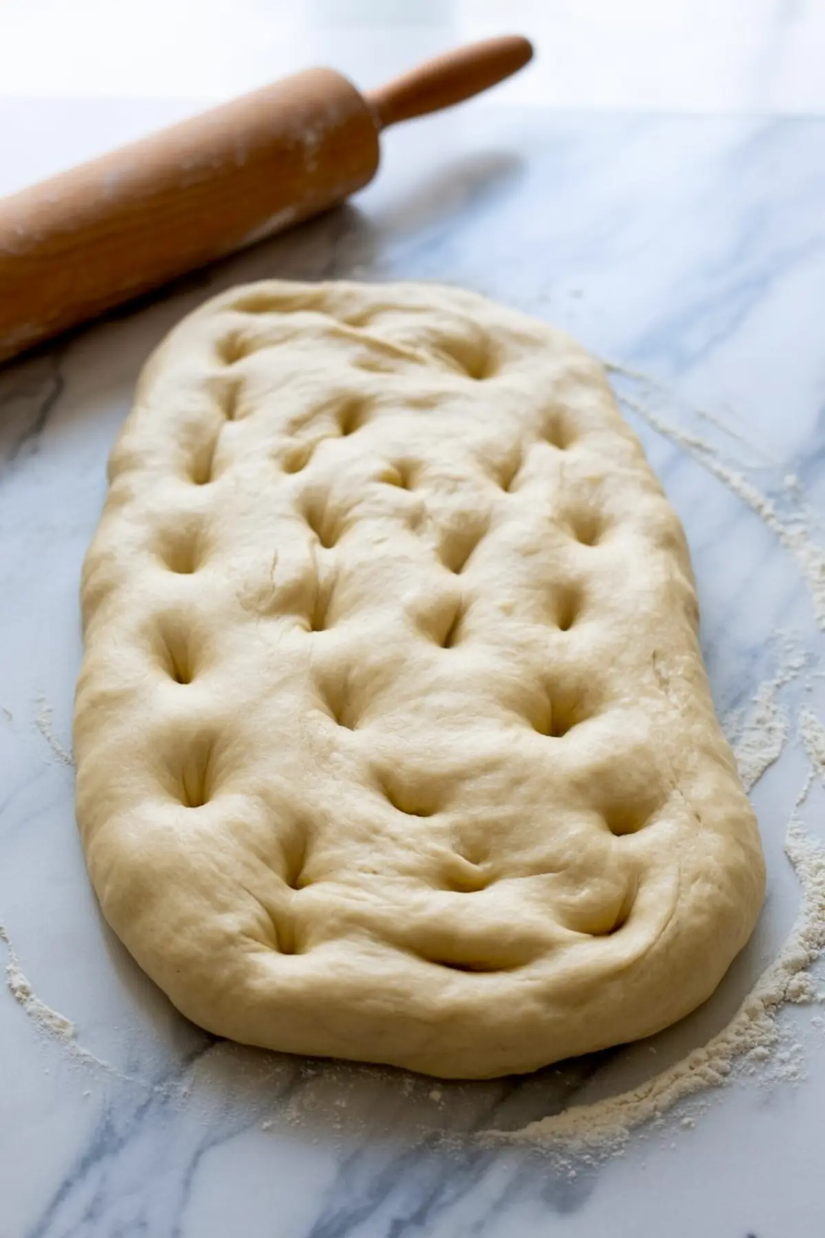 Unbaked focaccia dough shaped into a rectangle on a floured marble surface, with deep finger indentations and a wooden rolling pin placed nearby.

