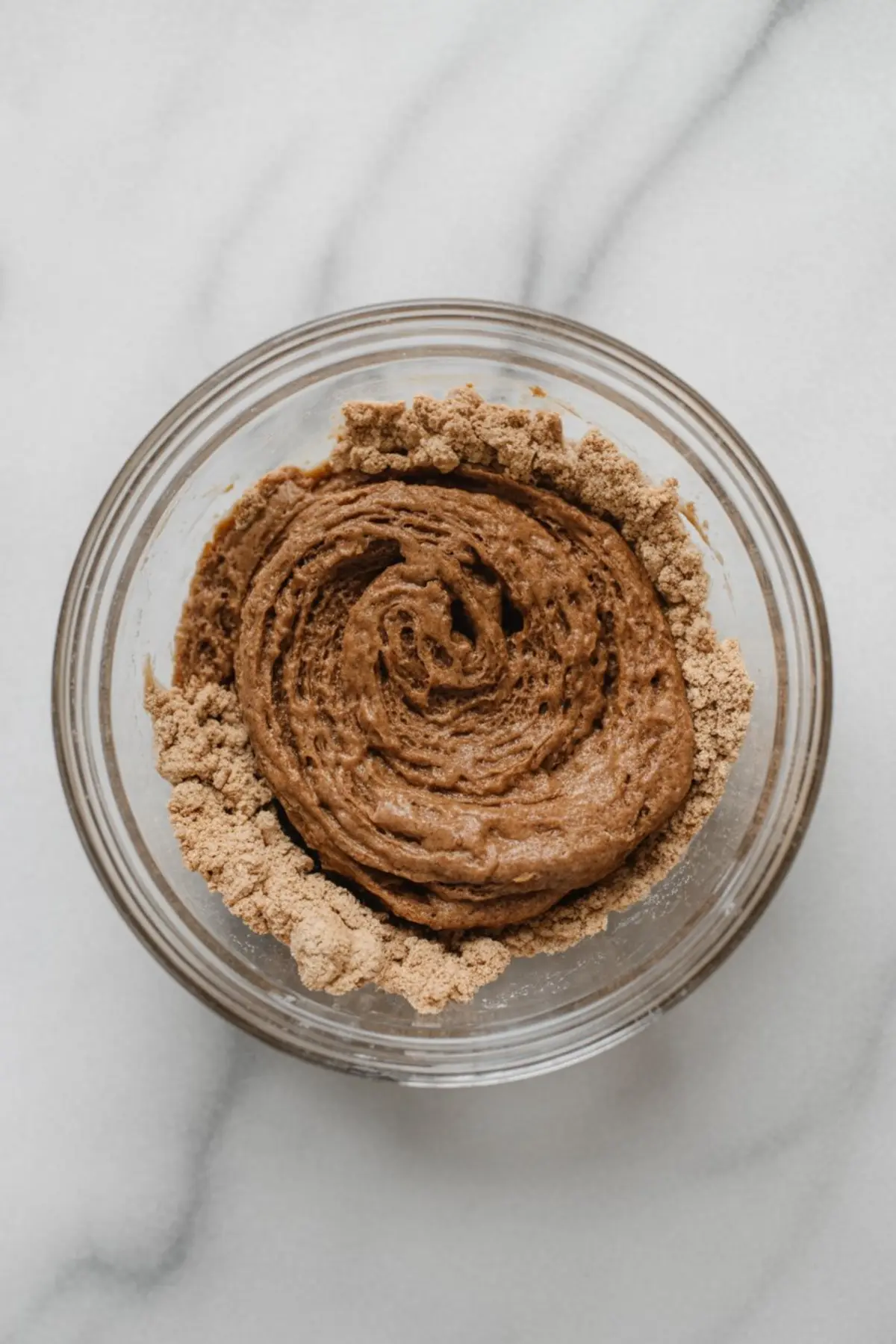 Glass bowl with creamy cinnamon filling mixture, partially blended with brown sugar, placed on a white marble surface.