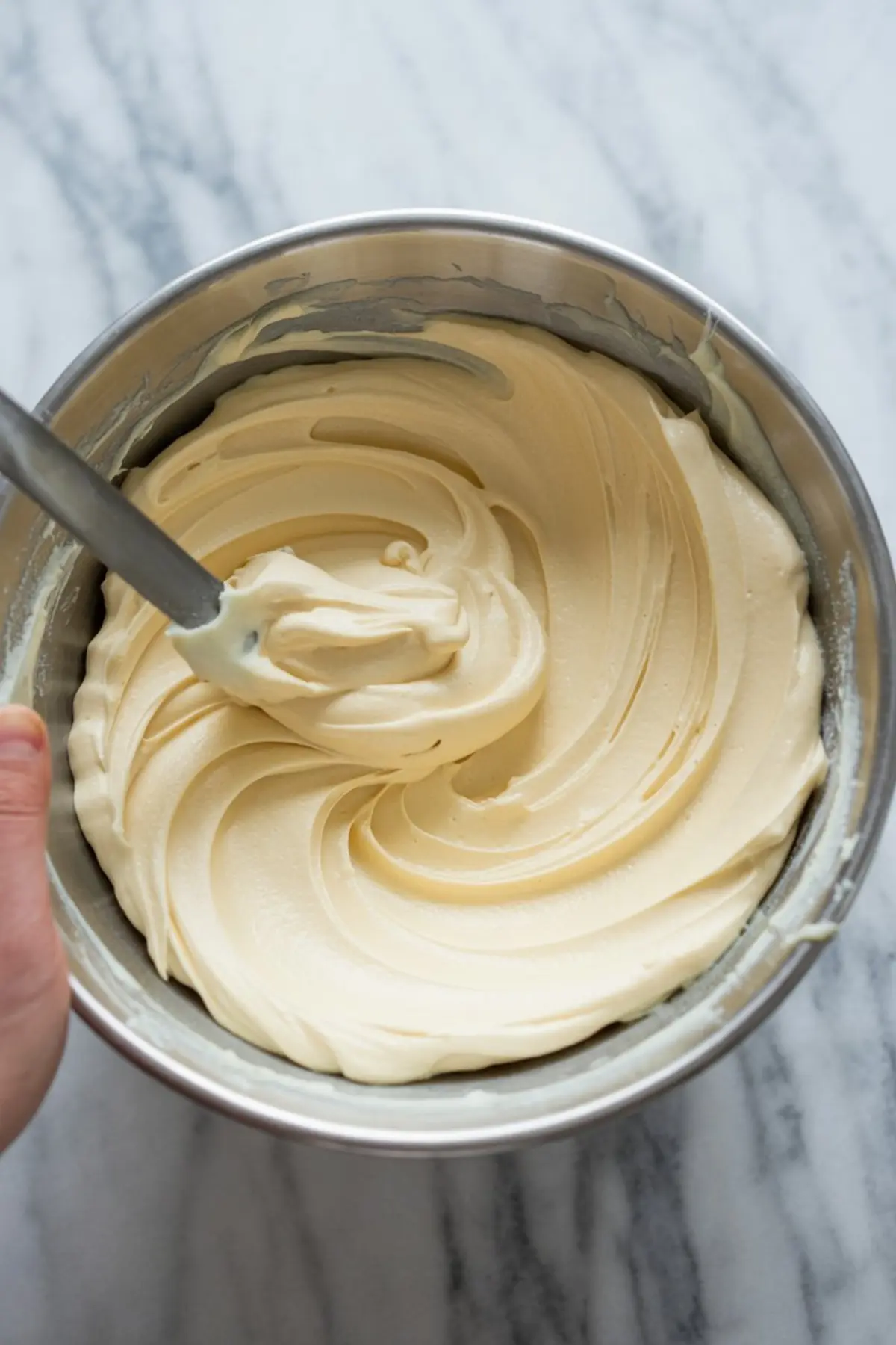 Creamy vanilla cake batter being mixed in a metal bowl with a rubber spatula, forming smooth swirls against a marble countertop.