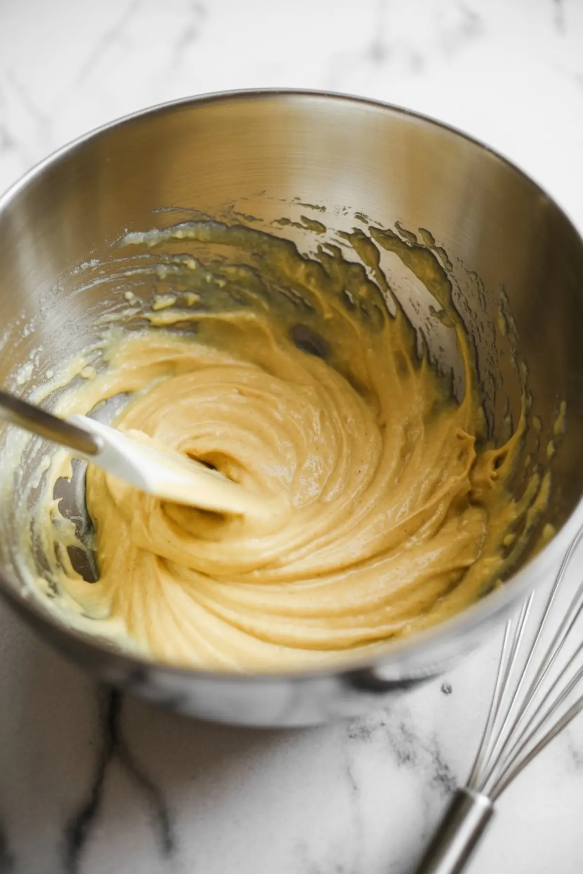 Golden cinnamon cake batter in a metal mixing bowl with a rubber spatula, showing a thick and glossy texture against a marble background.