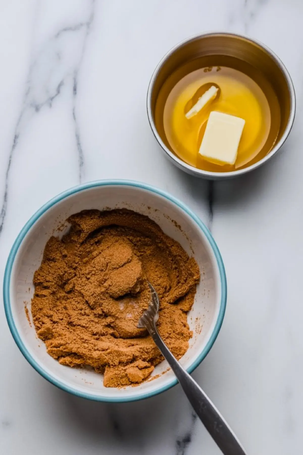 Bowl of cinnamon-sugar mixture with fork, next to small metal bowl containing melted butter and a pat of solid butter, on marble counter for cinnamon bread filling.