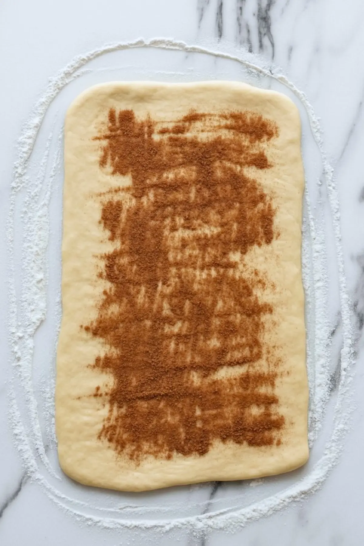 Rolled-out dough rectangle topped with brown cinnamon-sugar mixture, resting on floured marble surface, ready for rolling into cinnamon swirl loaf.
